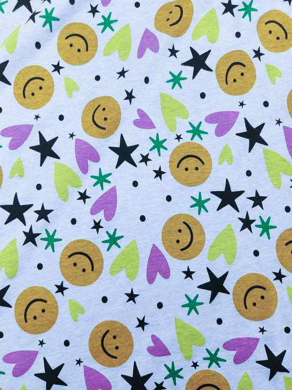 The Super Cheap Fabrics Printed Jersey - Hallow (165cm) features a playful design on lightweight poly/cotton fabric with yellow smiley faces, black stars, dots, and scattered green and purple hearts on a white background.