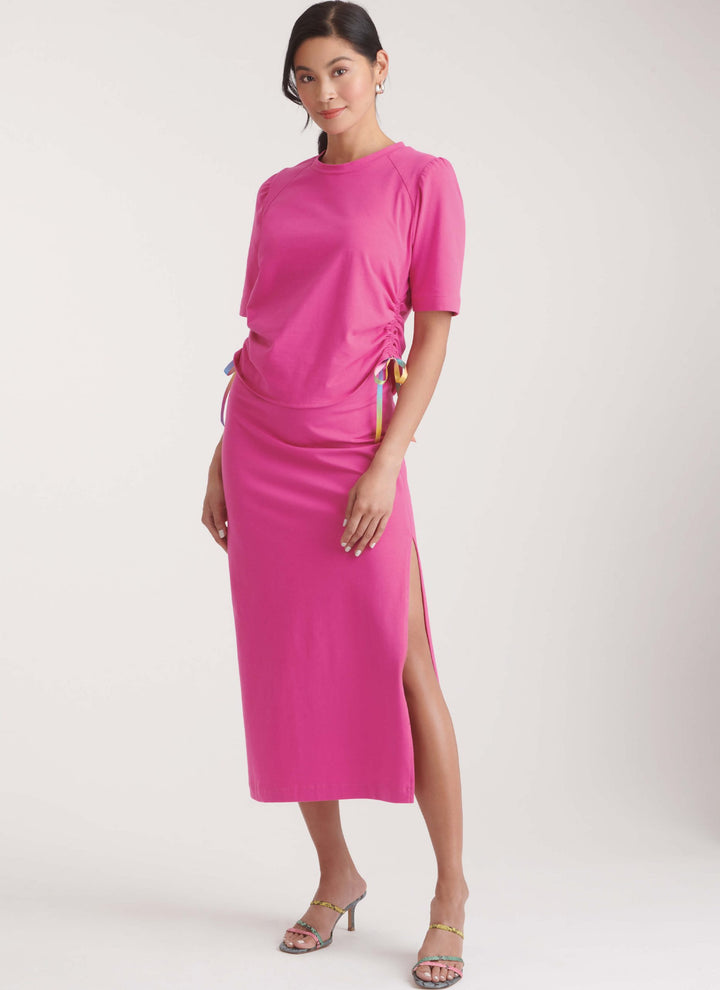 A woman poses in a pink midi skirt set, featuring side ruching and a high slit, made using the NEW LOOK 6782 Misses’ Knit Top and Skirt pattern from Super Cheap Fabrics. She pairs it with strappy heeled sandals against a plain background.