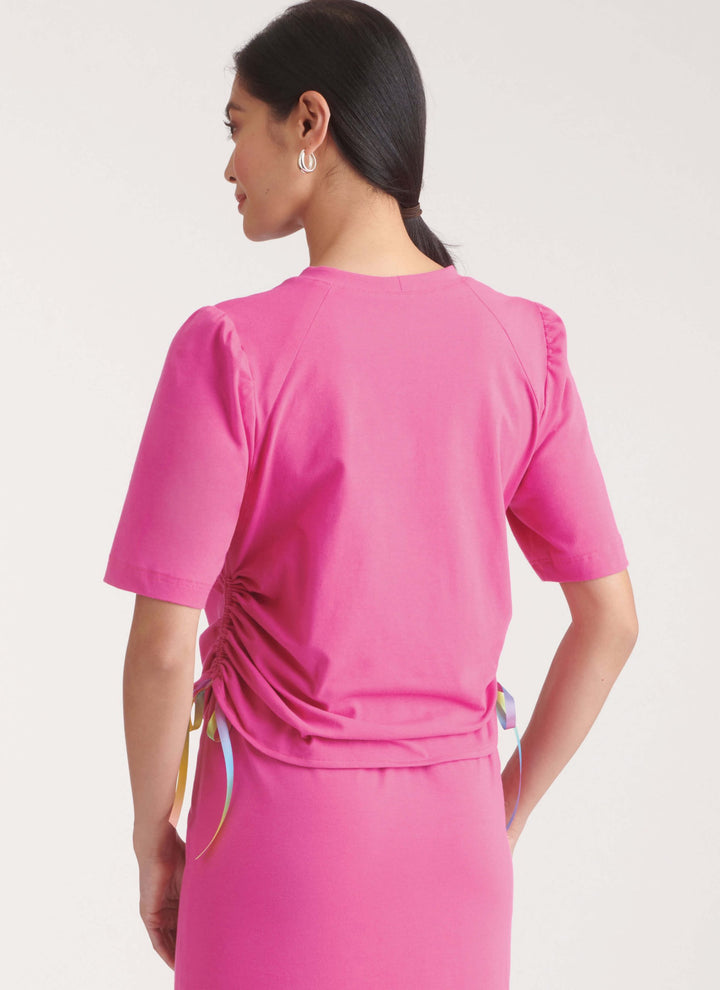 A woman with dark hair in a low ponytail models the Super Cheap Fabrics "Pattern - NEW LOOK - 6782 - Misses’ Knit Top and Skirt" set, featuring a bright pink ruched stretch top with rainbow ribbons and matching midi skirt, shown from the back.