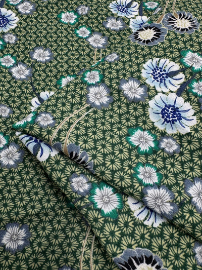 The Printed Crepe - Botan from Super Cheap Fabrics features elegant green fabric with detailed geometric patterns and floral designs in blue, white, and black. The flowers are intricate and scattered across the 148cm textile, which appears layered.