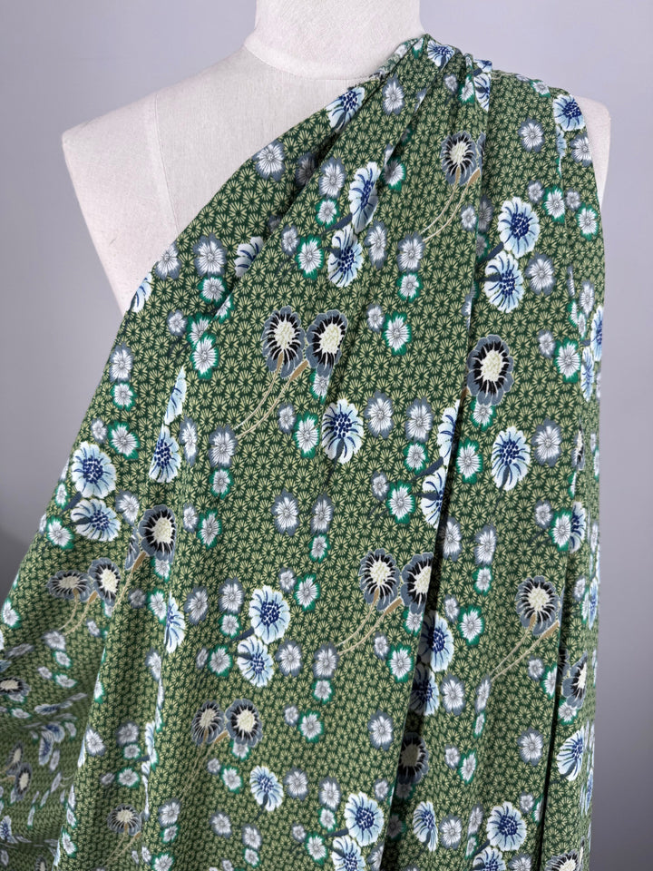 The Printed Crepe - Botan by Super Cheap Fabrics, draped over a mannequin, features an elegant floral pattern with blue, white, and black flowers surrounded by small green leaves on its 148cm wide green fabric.