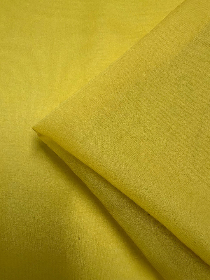 A close-up of a piece of Super Cheap Fabrics' Silk Georgette in Buttercup folded over itself. The lightweight, smooth fabric displays a slightly shiny texture, making it perfect for spring and summer outfits. The image primarily highlights the vibrant yellow color and delicate texture of this 135cm wide material.