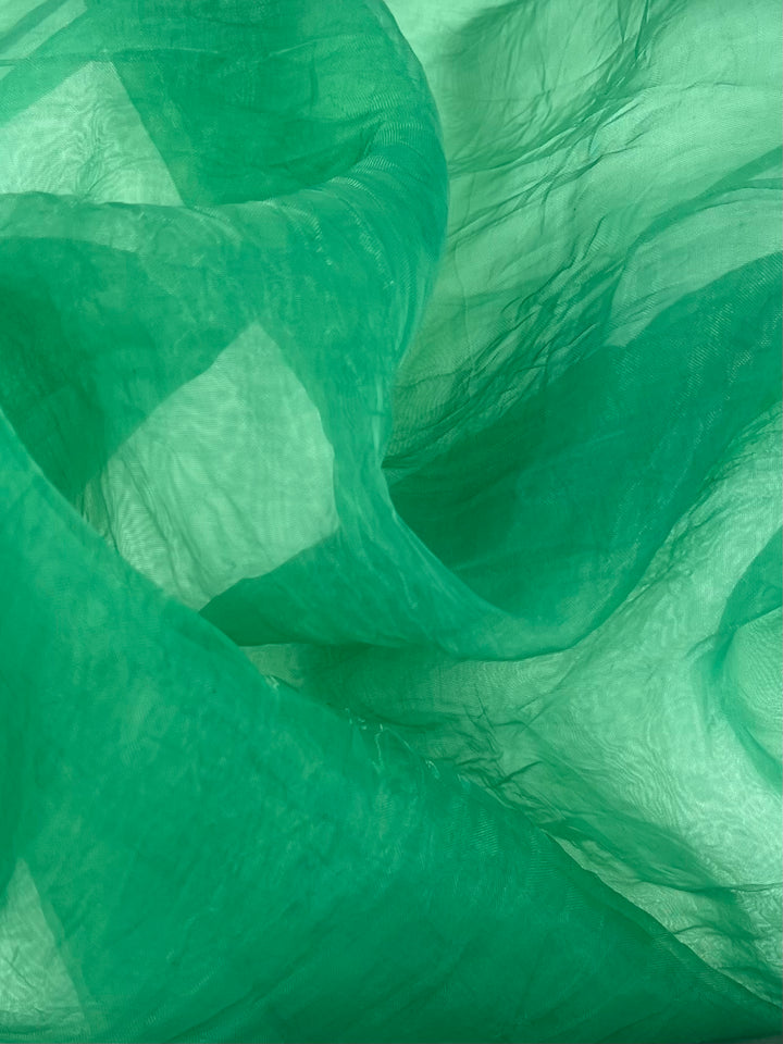 A close-up of flowing, translucent green Organza - Green Bee - 150cm from Super Cheap Fabrics with soft folds and ripples, creating abstract patterns and textures in varying shades of green. The light and shadow play across the 100% Nylon material, enhancing the sense of depth and movement ideal for tops and dresses.