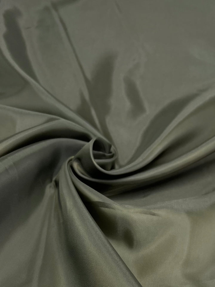A close-up image of a piece of shiny, smooth, olive-green lightweight fabric. The material is gently gathered and twisted in the center, creating soft folds and highlights that catch the light, giving the Super Cheap Fabrics Lining - Olive - 112cm a glossy appearance.