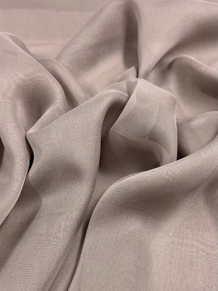 A close-up image showcases the Silk Georgette - Quail - 135cm from Super Cheap Fabrics, highlighting its soft, beige hue with a smooth and slightly sheer texture. The fabric is loosely draped and folded, creating gentle curves and shadows, making it ideal for elegant summer outfits due to its delicate and lightweight nature.