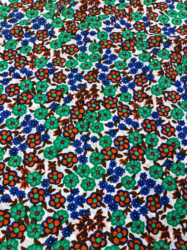 The Salt Shrunk Print - Kaleid from Super Cheap Fabrics features a lively floral pattern with green, orange, and blue flowers on an elegant white background. The vibrant, densely packed design ensures a colorful appearance that captures attention effortlessly.