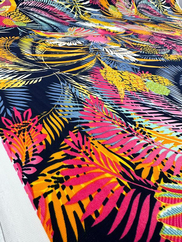 Introducing the Printed Lycra - Jumba - 150cm by Super Cheap Fabrics: A striking, vibrant fabric that boasts a bold tropical leaf pattern with large, colorful leaves in shades of pink, yellow, orange, and green on a dark background. This beautiful print creates an energetic visual perfect for making a statement.