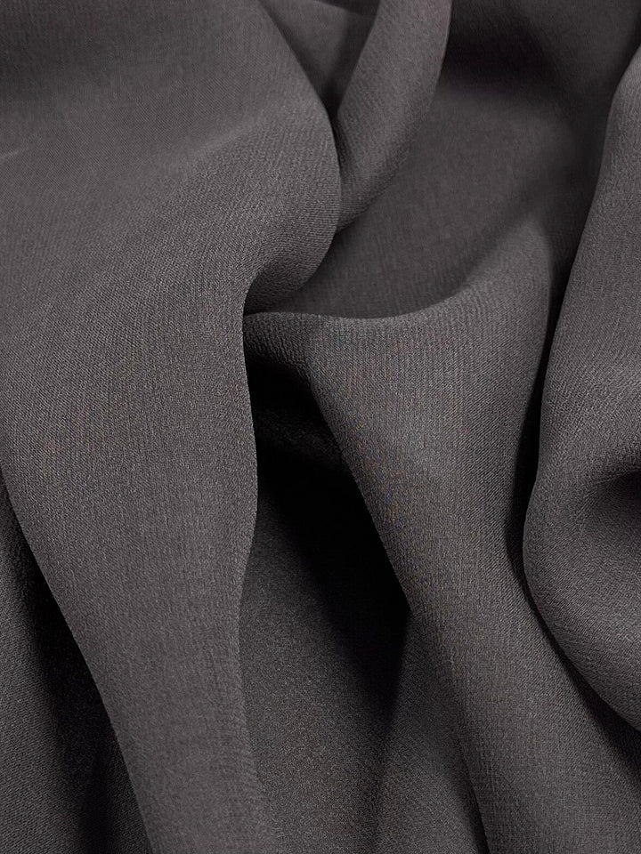 A close-up of Super Cheap Fabrics' "Silk Georgette - Pewter - 135cm" showcases its dark gray hue with gentle folds and a soft texture, creating a flowing and smooth appearance. The 100% silk fabric appears lightweight and finely woven, with subtle shadows and highlights that add depth to the image.