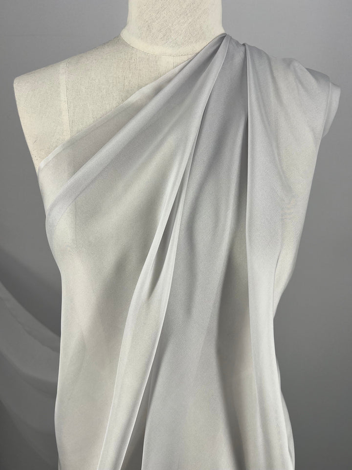 A white, translucent, extra light weight fabric from Super Cheap Fabrics' Pure Silk Chiffon - Silver collection is elegantly draped over a dress form, covering one shoulder and cascading diagonally across the torso. Made of 100% silk, its bridal blush undertones are enhanced by the subtle gray background, which amplifies the simplicity and elegance of the setup. This luxurious fabric measures 140cm in width.