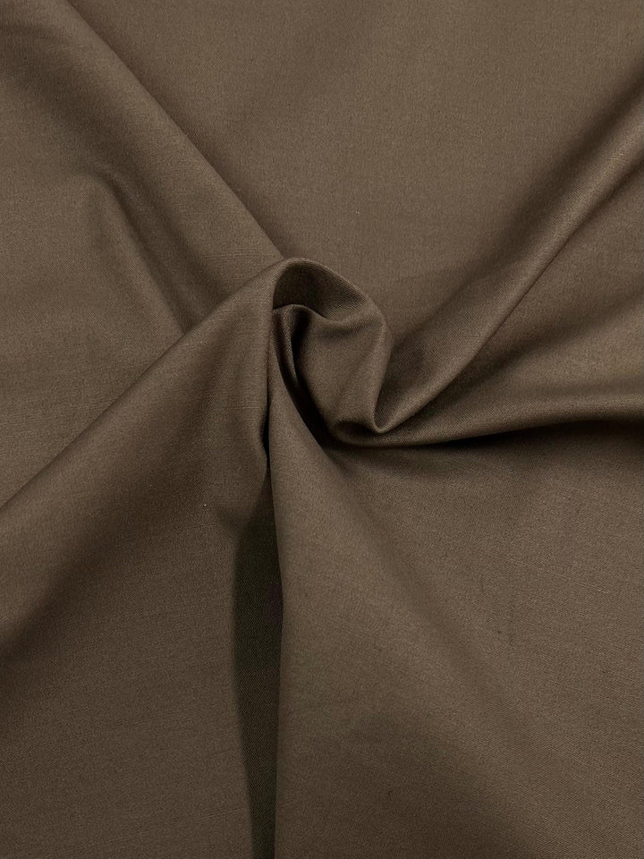 A close-up image of a brown, medium-heavy weight fabric with a subtle sheen. This 100% cotton material is gathered and twisted in the center, creating soft folds and creases that add texture to its smooth surface, ideal for workwear. The product is Cotton Sateen - Kangaroo - 118cm by Super Cheap Fabrics.