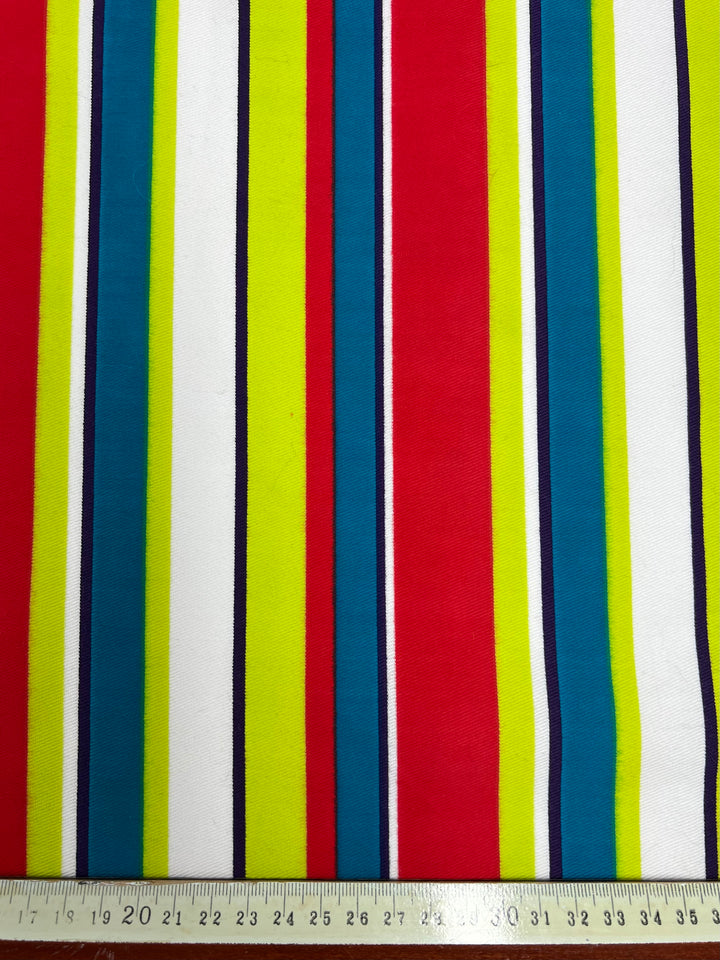 The Printed Denim - Temperature Stripes by Super Cheap Fabrics is a colorful, light medium weight 100% cotton fabric featuring vertical stripes in various colors, including red, yellow, green, blue, black, and white. A measuring tape along the bottom edge shows the length in centimeters from 17 to 35. The stripe widths vary for added visual interest.